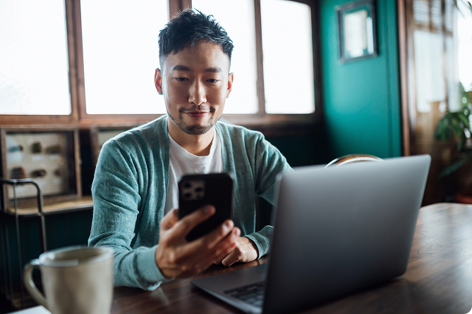 A man looking at his smartphone with an open laptop in front of him at a wooden table.