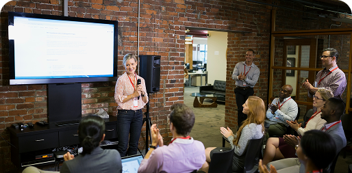 A woman giving a presentation to an audience in an office with exposed brick walls and a large screen.